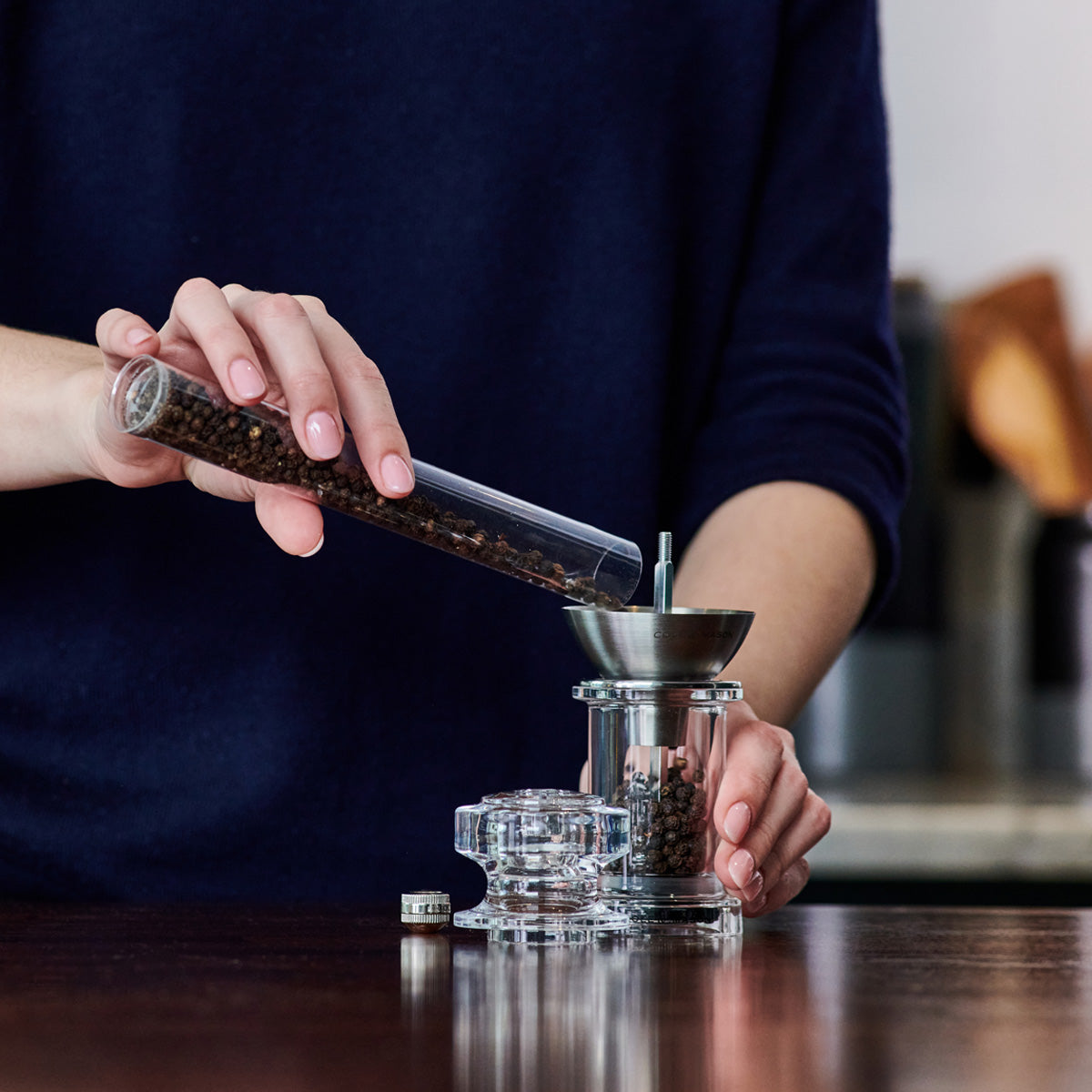 A person pours whole peppercorns from a glass tube into a transparent pepper grinder with a metal funnel on a dark kitchen counter. Kitchen items are blurred in the background.