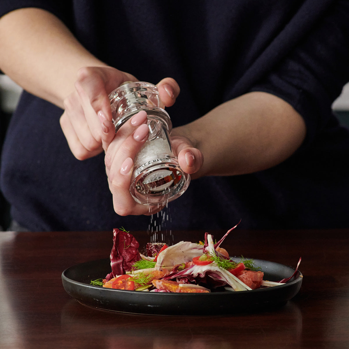 A person sprinkles salt from a clear grinder onto a colorful salad with greens, tomatoes, and red leaves on a black plate.
