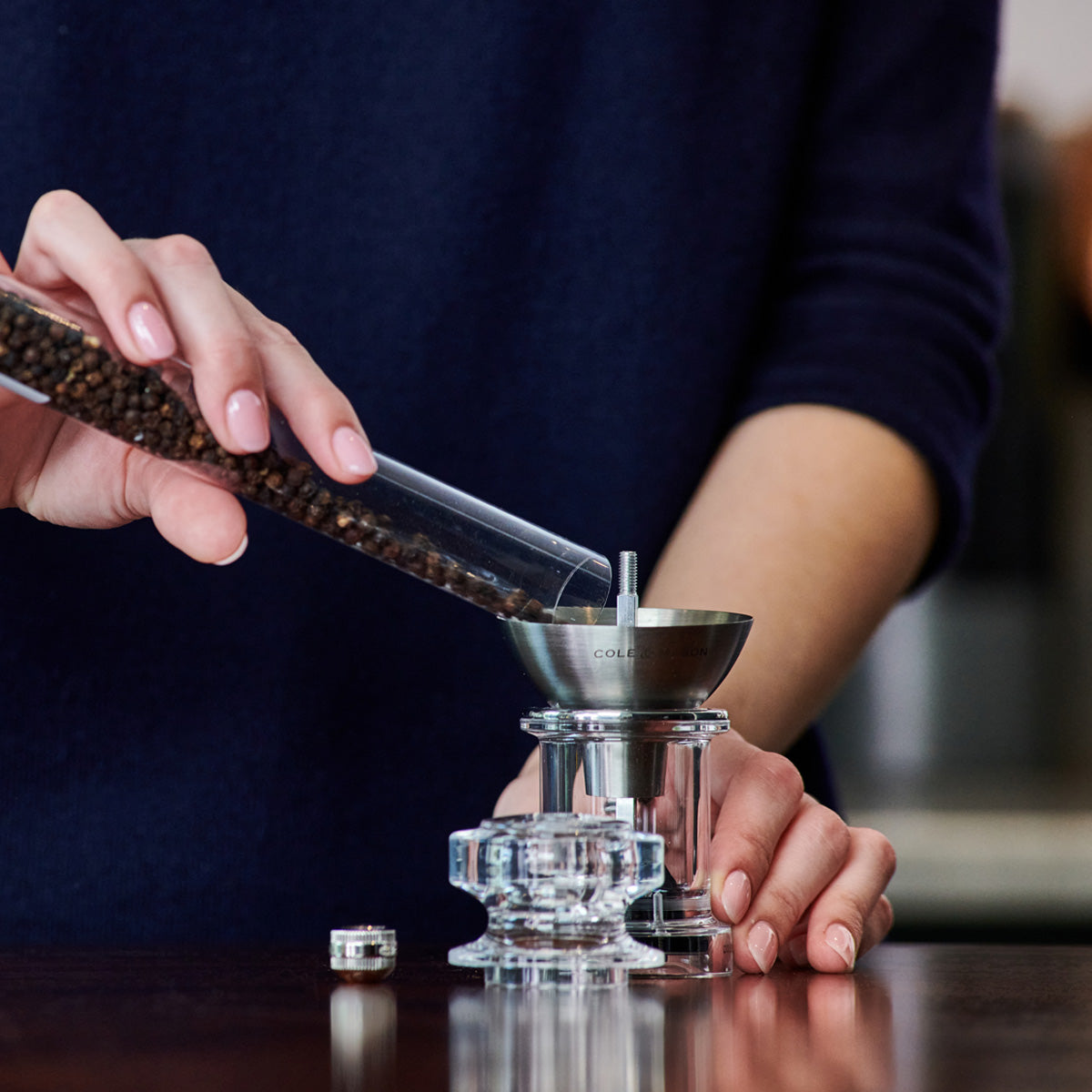 A person pours whole black peppercorns from a glass tube into the top of a transparent pepper grinder on a dark wooden surface.