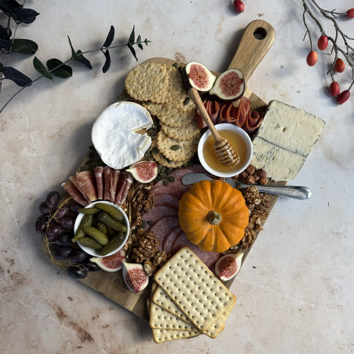 A charcuterie board with crackers, brie, blue cheese, salami, figs, prosciutto, grapes, cornichons, walnuts, honey, and a small pumpkin on a wooden board, surrounded by autumn leaves and branches.