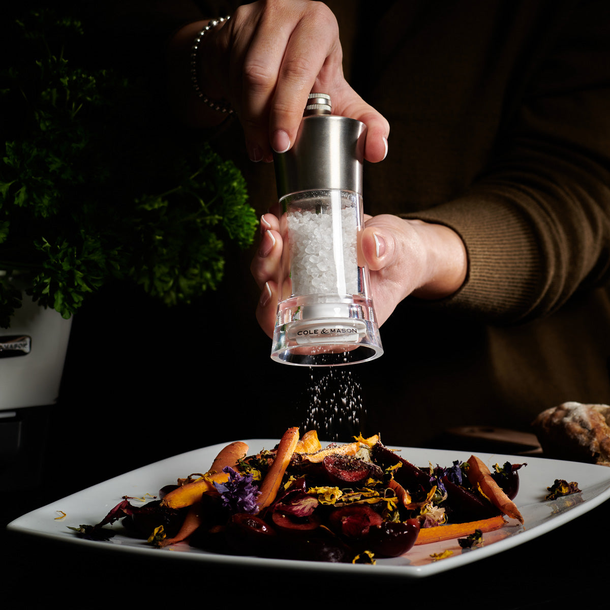 A person grinds sea salt over a plated dish of colorful roasted vegetables and edible flowers, with fresh herbs visible in the background.