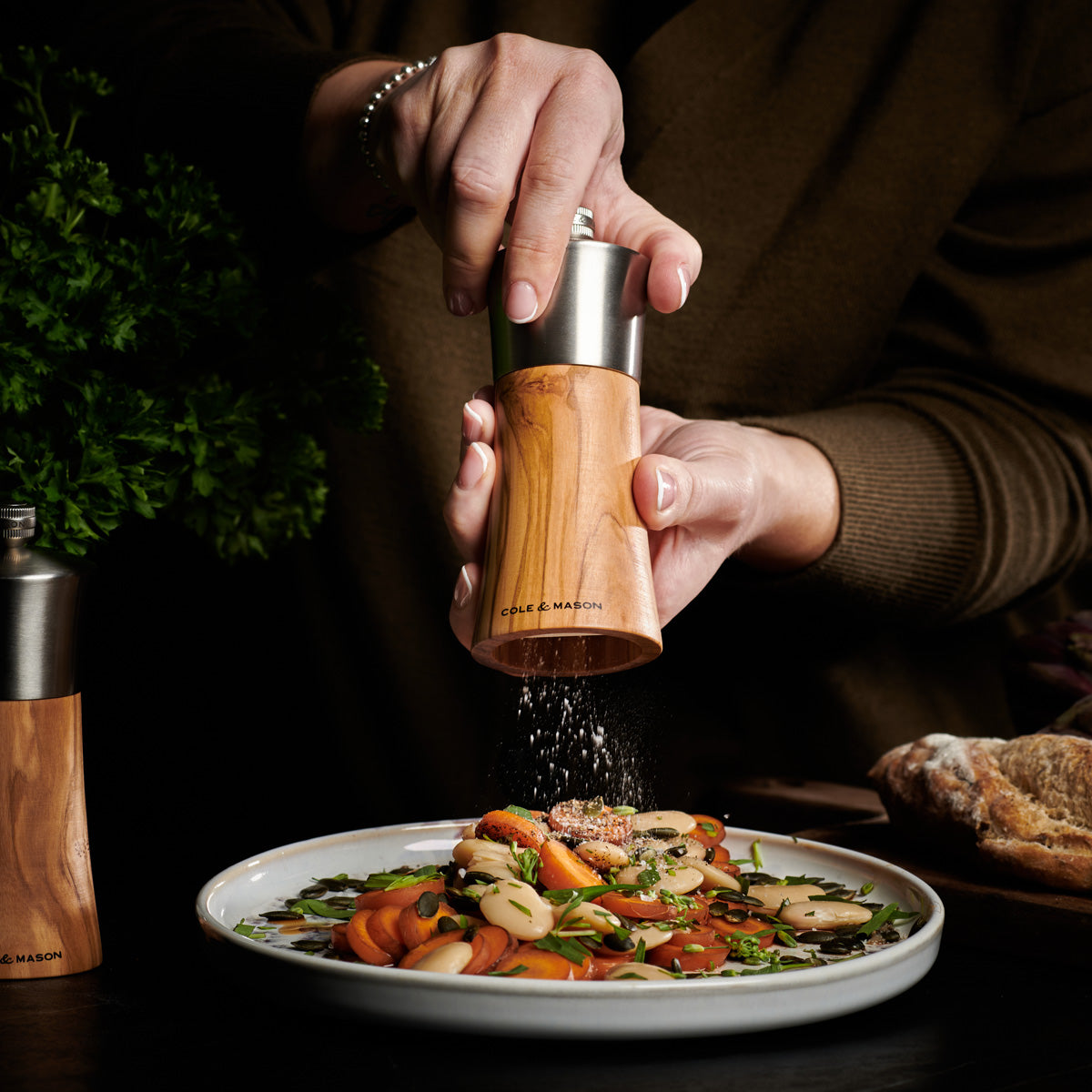 A person grinds pepper over a plate of salad with sliced vegetables and beans, using a wooden pepper mill. Fresh herbs and bread are visible in the background.
