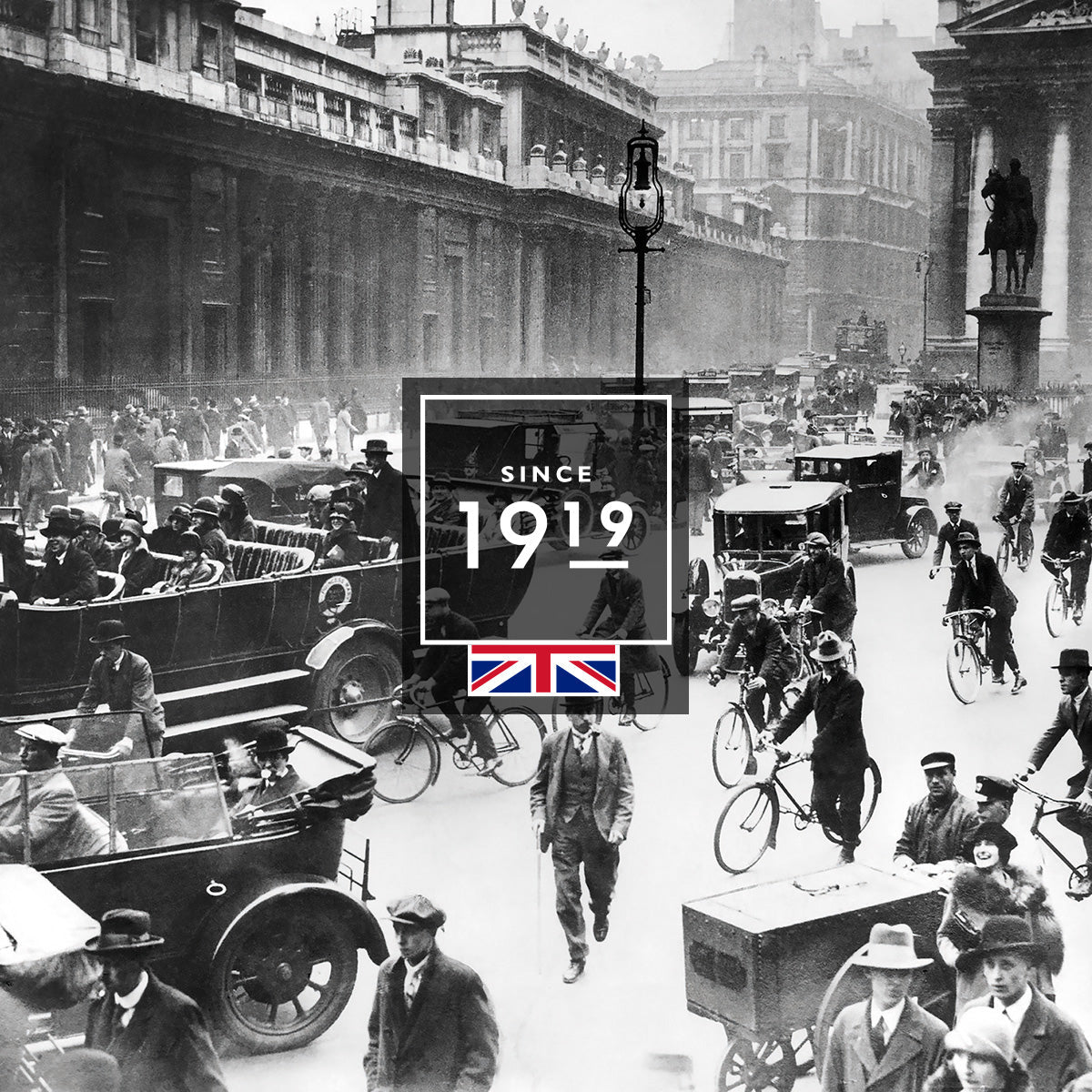 A busy black-and-white street scene from 1919 in the UK, showing people walking, cycling, and riding in early cars. A Since 1919 graphic with a British flag is centered over the image.