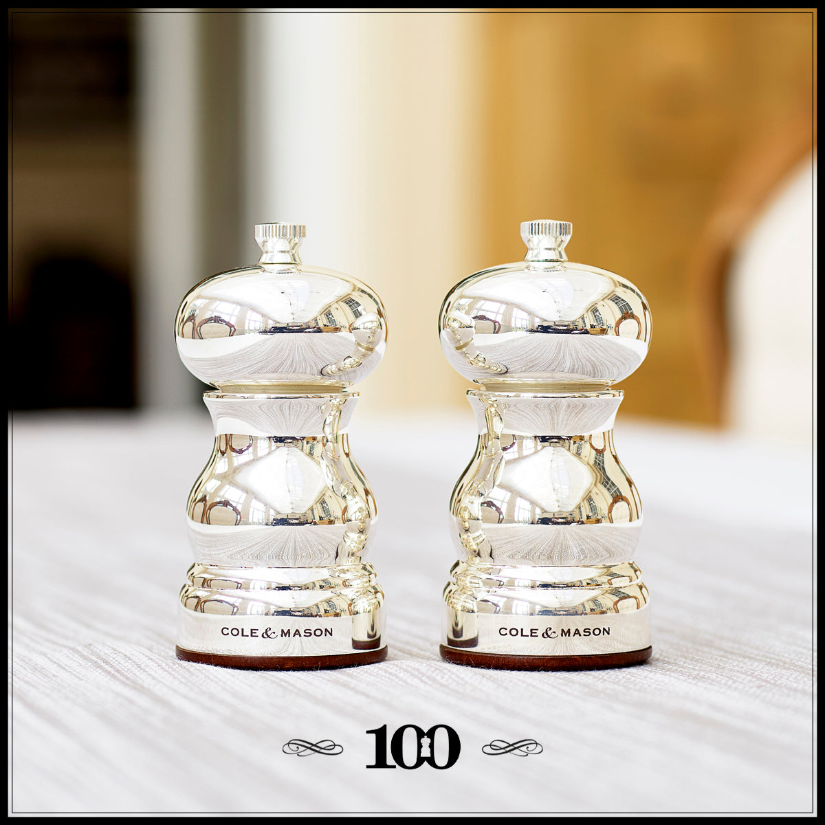 Two shiny silver Cole & Mason salt and pepper mills are placed side by side on a white surface, with the number 100 printed below them in black. The background is softly blurred.