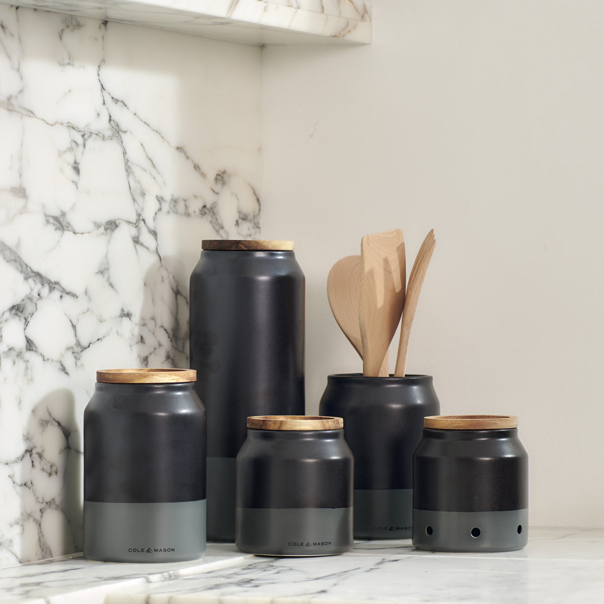 Five black and gray kitchen canisters with wooden lids are arranged on a marble countertop. One canister holds wooden utensils; the background features a marble backsplash and cream-colored wall.