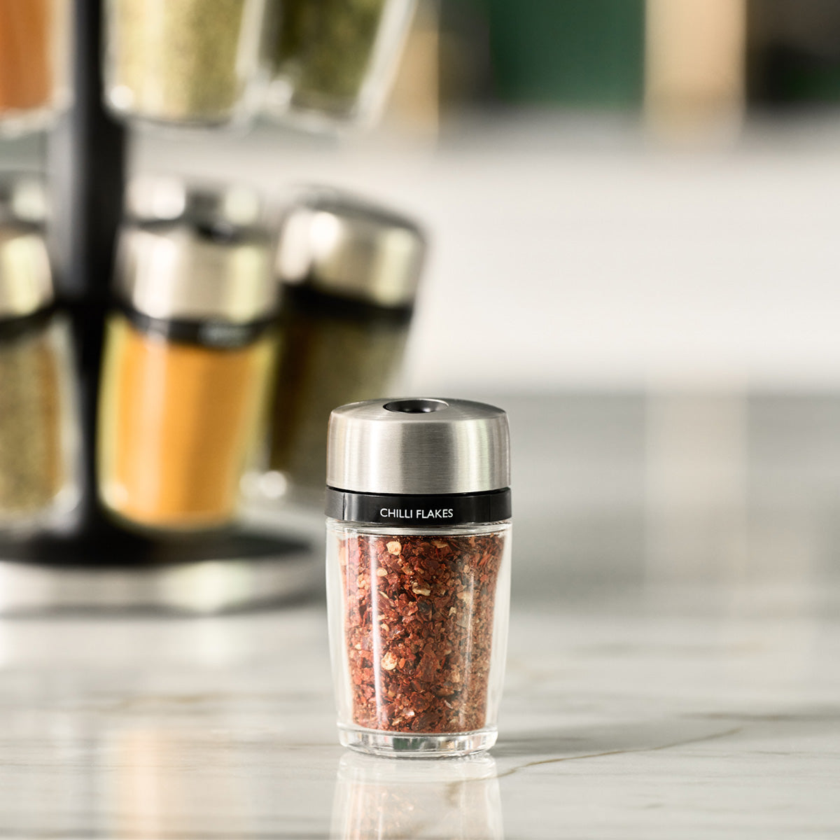 A glass jar labeled Chili Flakes filled with red chili flakes sits on a marble countertop, with a spice rack holding other jars visible in the blurred background.