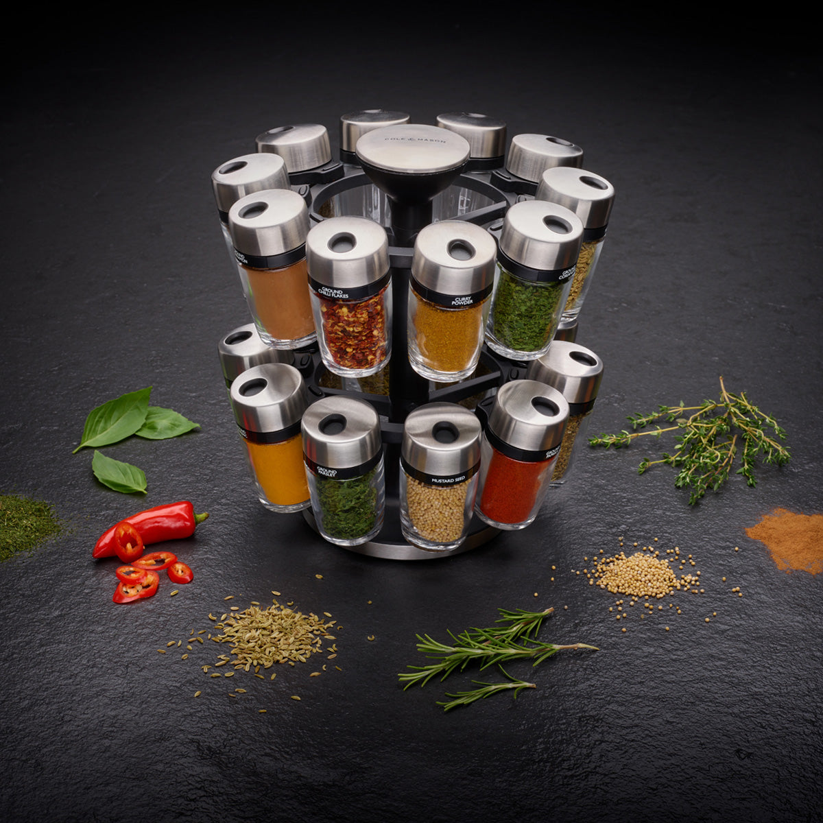 A rotating spice rack with labeled glass jars filled with various colorful spices is surrounded by fresh herbs, chili peppers, and piles of loose spices on a dark surface.