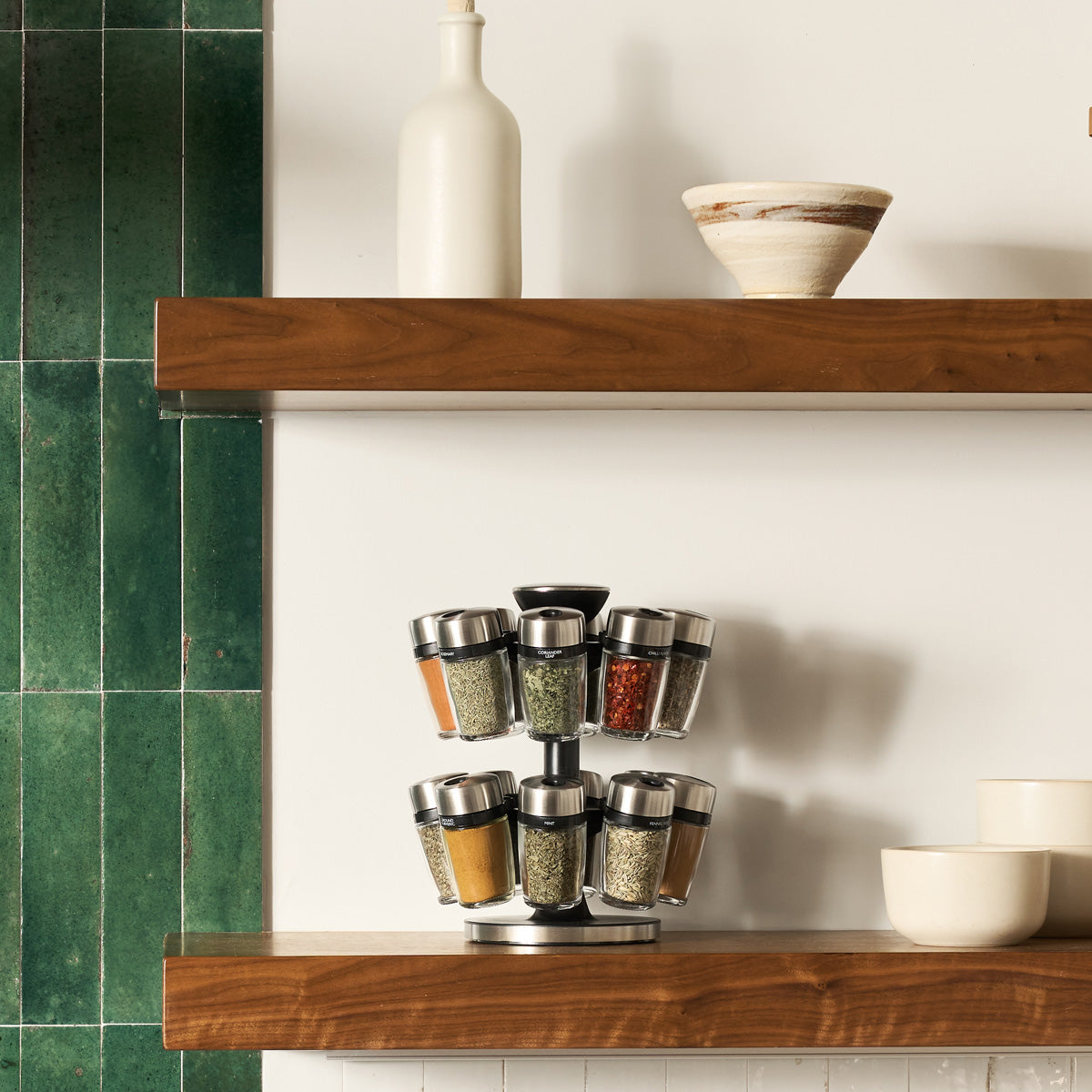 A modern kitchen with wooden shelves displaying ceramic bowls, a bottle, and a rotating spice rack filled with assorted spices, set against a green tiled and white wall background.