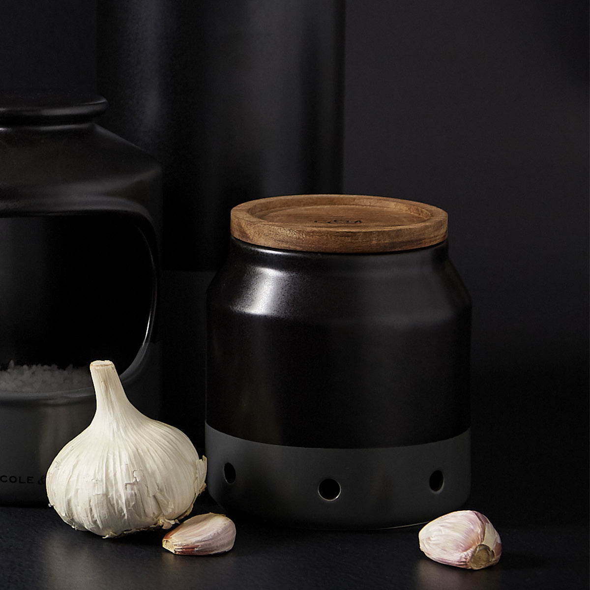 A black ceramic container with a wooden lid, featuring small holes near the base, sits on a dark surface beside a whole head of garlic and two loose garlic cloves.