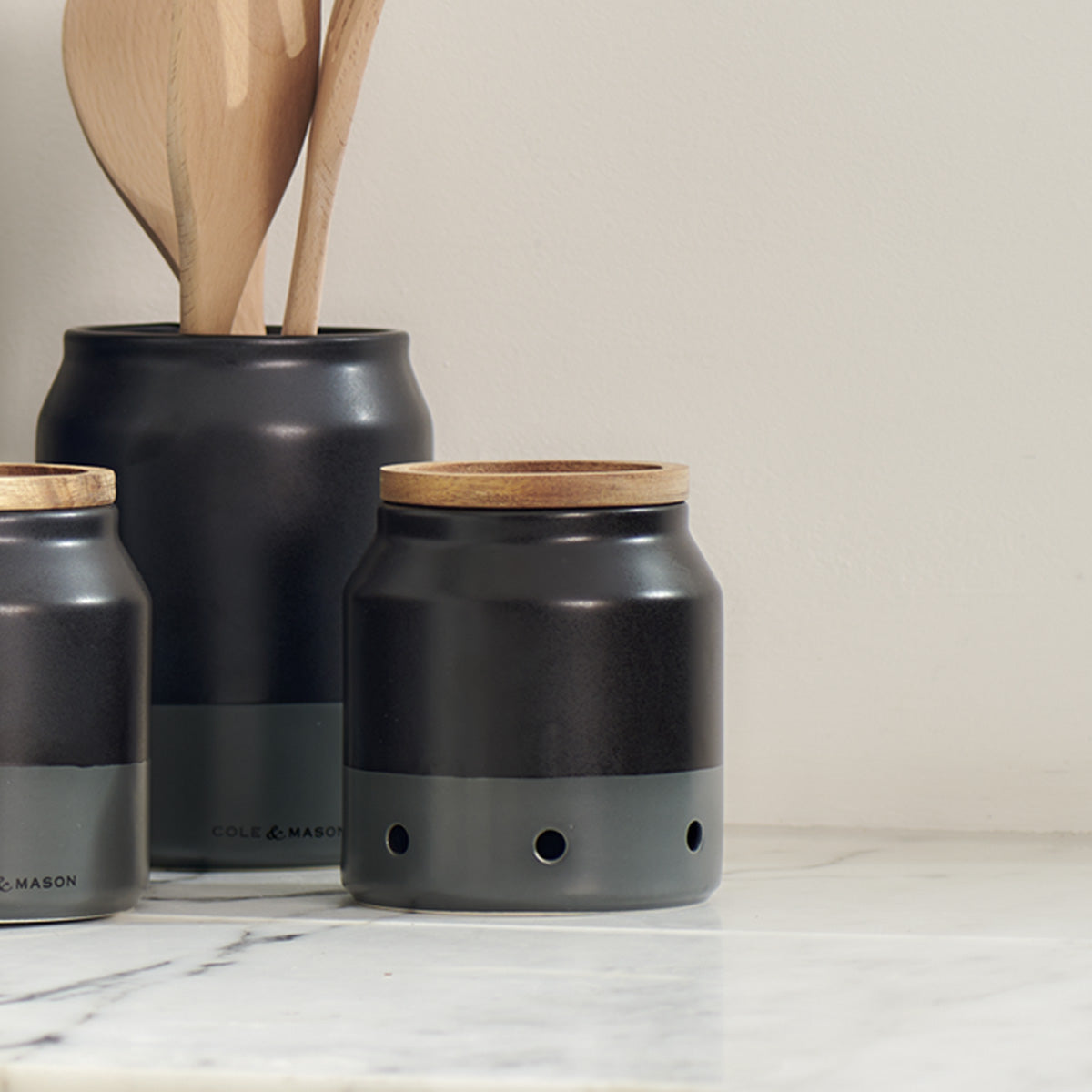 Three ceramic kitchen canisters with wooden lids and a utensil holder containing wooden spoons are arranged on a white marble countertop against a light-colored wall.