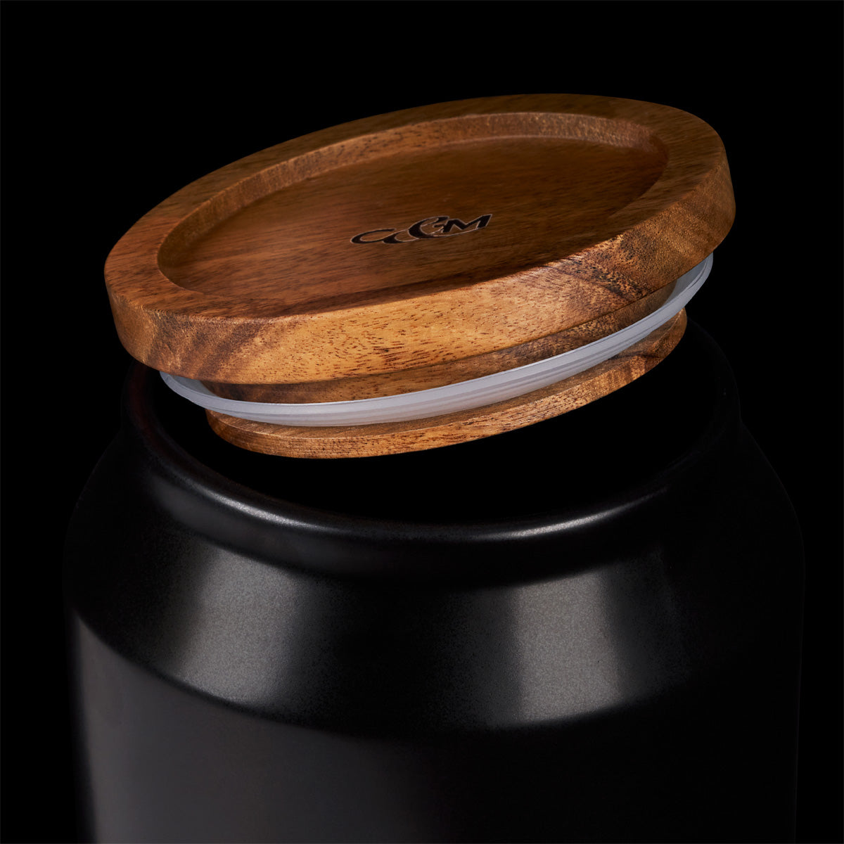 A close-up of a matte black container with a wooden lid, partially open, showing a white silicone sealing ring. The lid has a C&M logo engraved on top. The background is completely black.