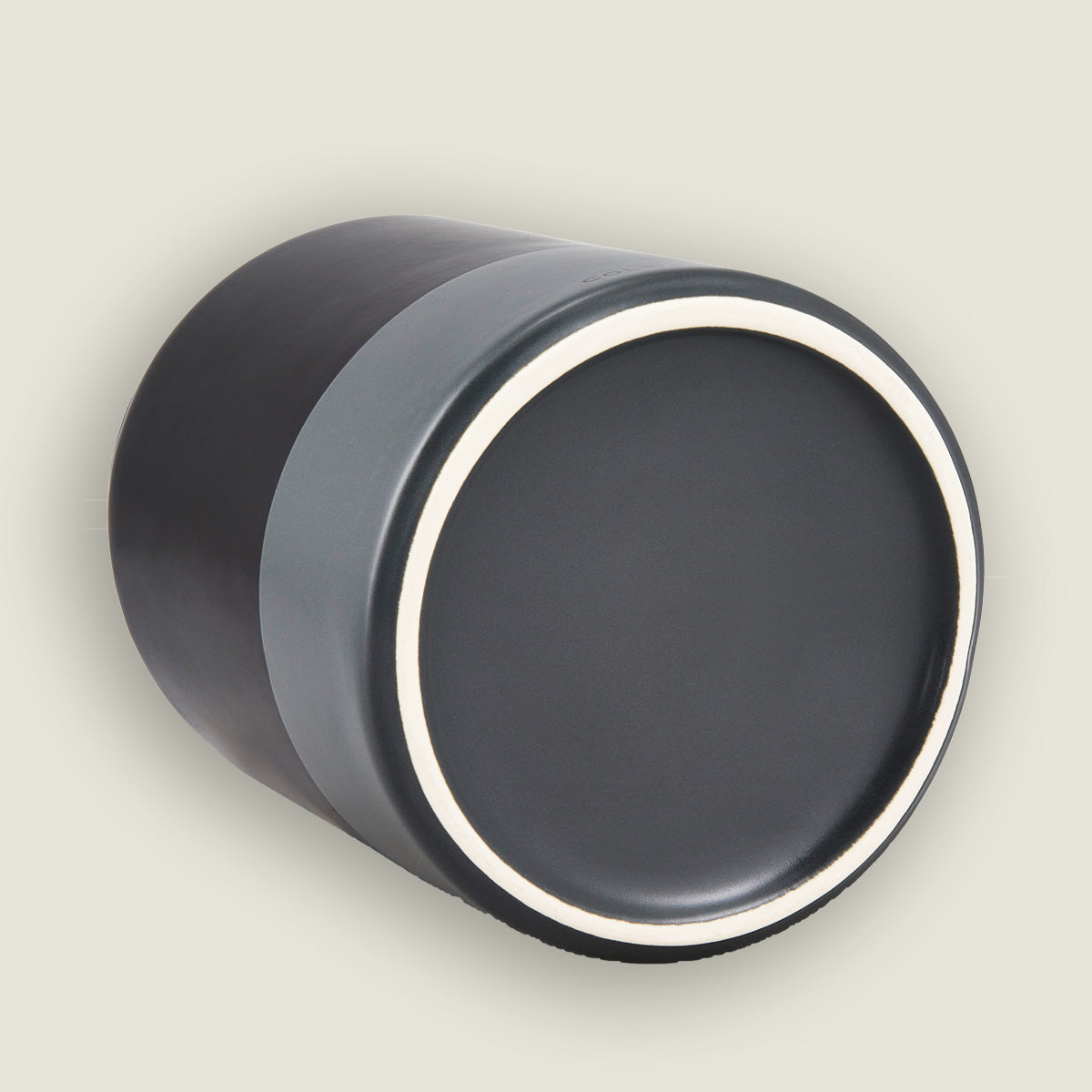 A cylindrical black and gray ceramic cup is lying on its side, showing a smooth, flat bottom rimmed with a thin white line.