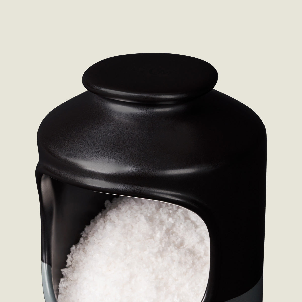 A close-up of a black ceramic container filled with coarse white salt, set against a light beige background. The container has a wide opening in the front and a smooth, rounded lid on top.