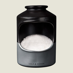 A ceramic salt cellar with a wide opening, filled with coarse sea salt. The container is black and gray with Cole & Mason printed on the front.