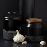 A black ceramic salt cellar filled with coarse salt, a matching black canister with a wooden lid, and a whole garlic bulb with two garlic cloves are arranged against a dark background.