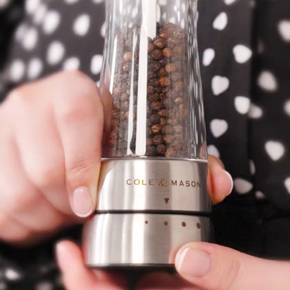 A person holds a Cole & Mason pepper grinder filled with peppercorns, adjusting the grind setting. The background shows a black shirt with white heart patterns.