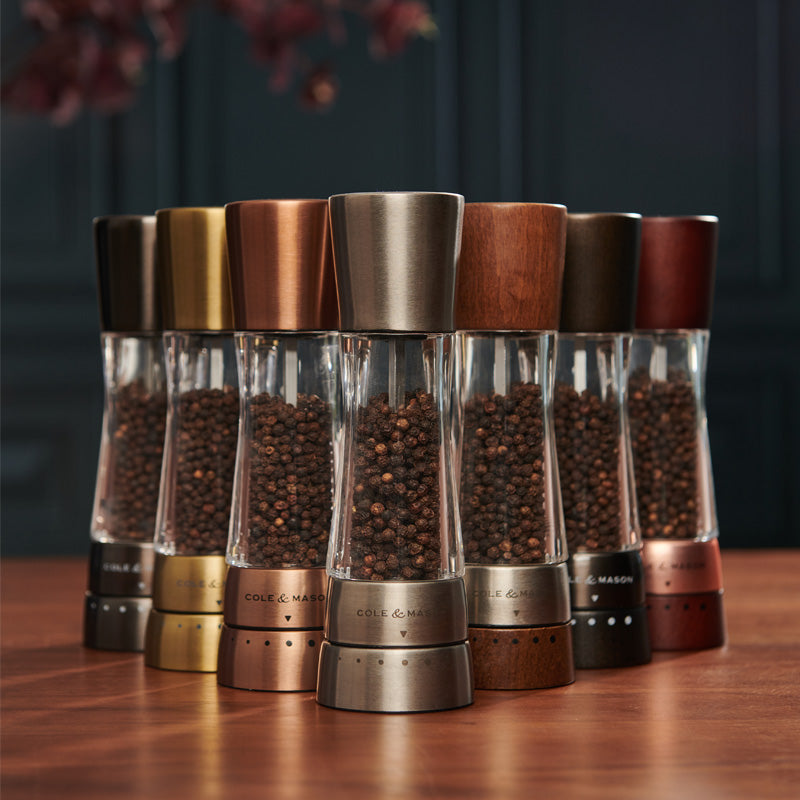 A row of seven pepper grinders filled with peppercorns, each with a different metallic or wooden finish, arranged on a wooden surface against a dark, blurred background.
