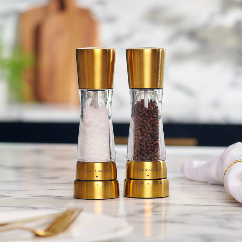 Two modern salt and pepper grinders with gold accents sit side by side on a marble countertop; one contains white salt crystals and the other black peppercorns. A napkin and blurred kitchen items are in the background.