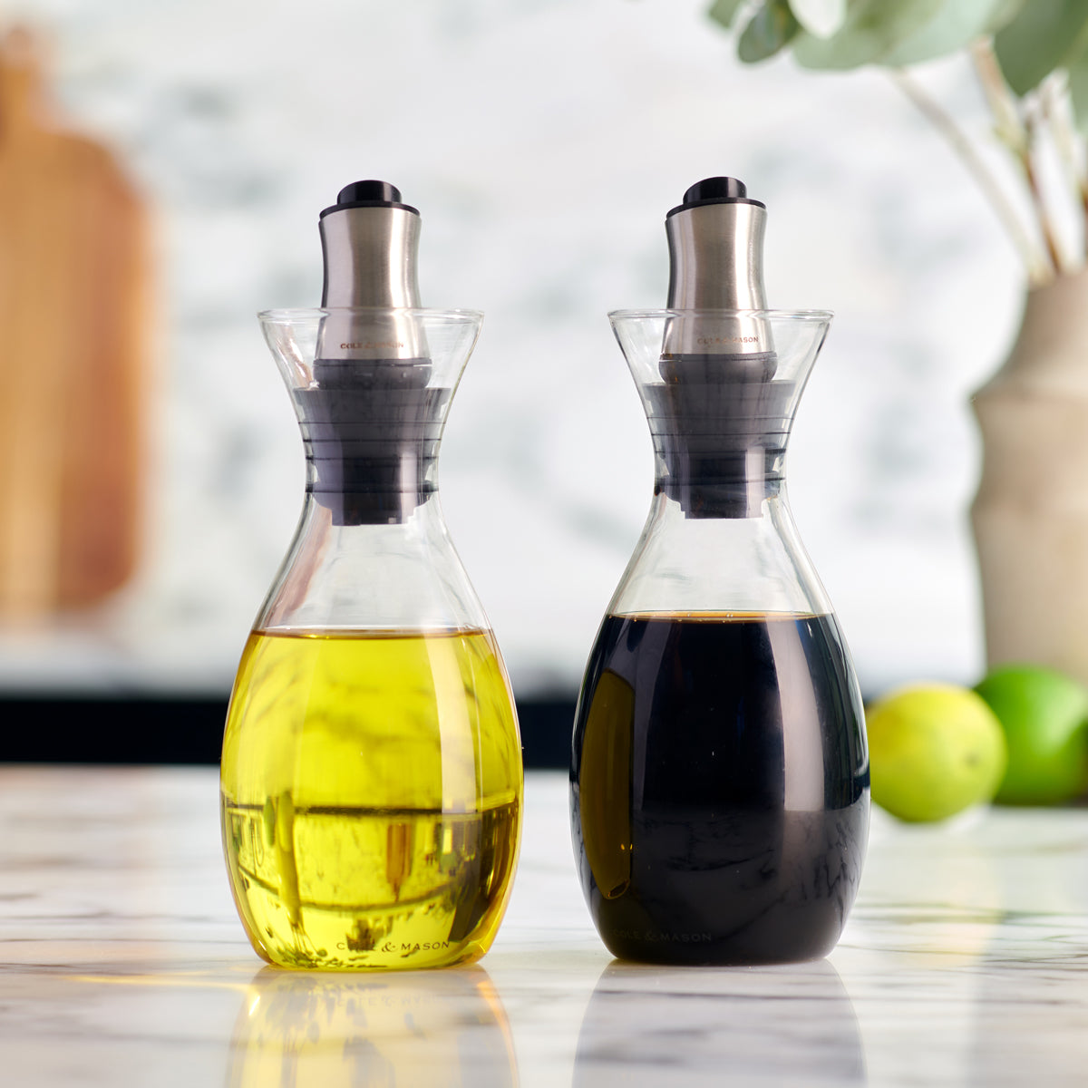 Two glass dispensers on a marble countertop, one filled with yellow olive oil and the other with dark balsamic vinegar. Both have metal and black plastic pour spouts. Blurred background with fruit and a vase.