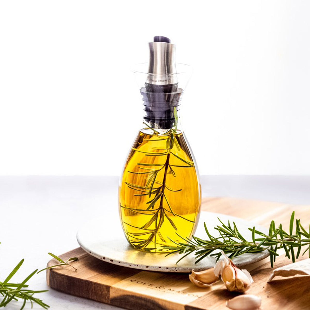 A glass bottle of olive oil infused with a sprig of rosemary sits on a plate atop a wooden board, surrounded by fresh rosemary and garlic cloves.
