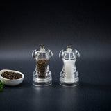Two clear Cole & Mason Acrylic Capstan Mills—one with black peppercorns, one with white salt—stand side by side on a dark surface. A small bowl of peppercorns completes this perfect Acrylic Capstan Mill Christmas Gift Hamper.
