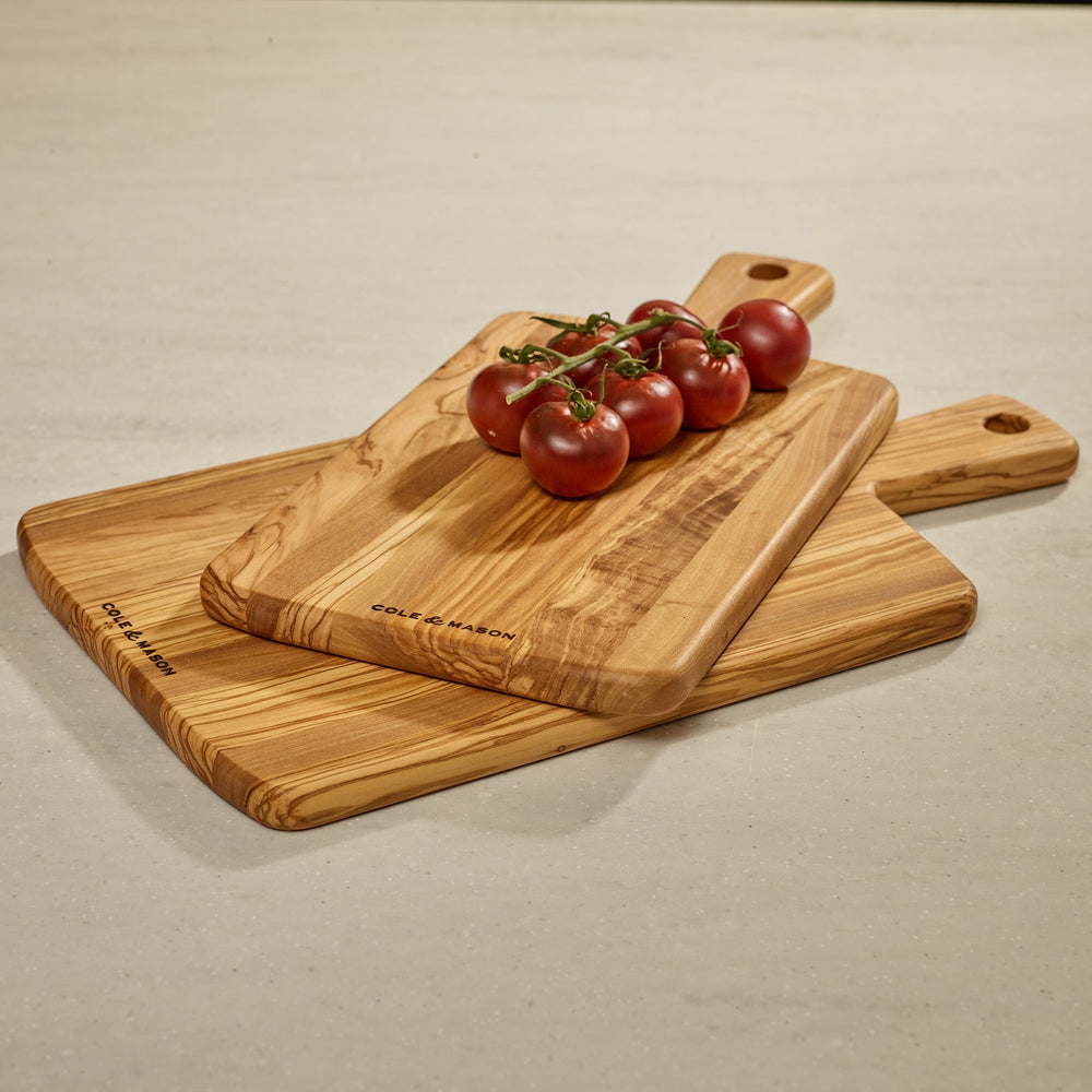 The Cole & Mason Portobello Olive Wood Serving Board, with a handle and hanging hole, showcases an elegant natural grain—ideal as a stylish antipasti board and shown upright on a light background.
