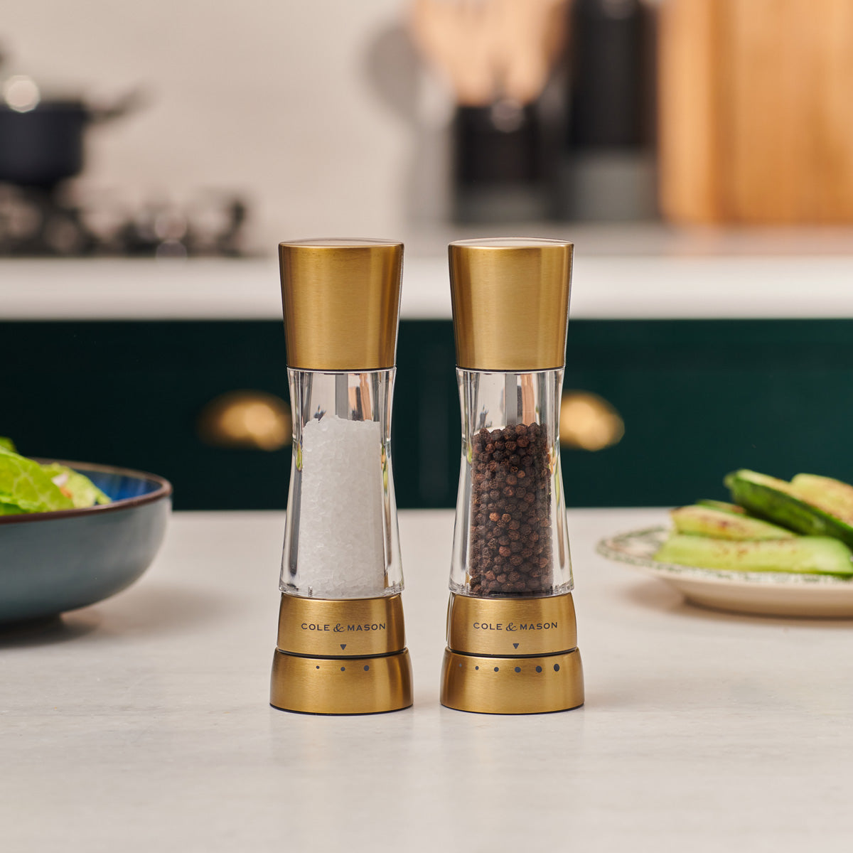 Two gold and clear Cole & Mason salt and pepper grinders are placed side by side on a kitchen counter, with a salad and vegetables in the background.