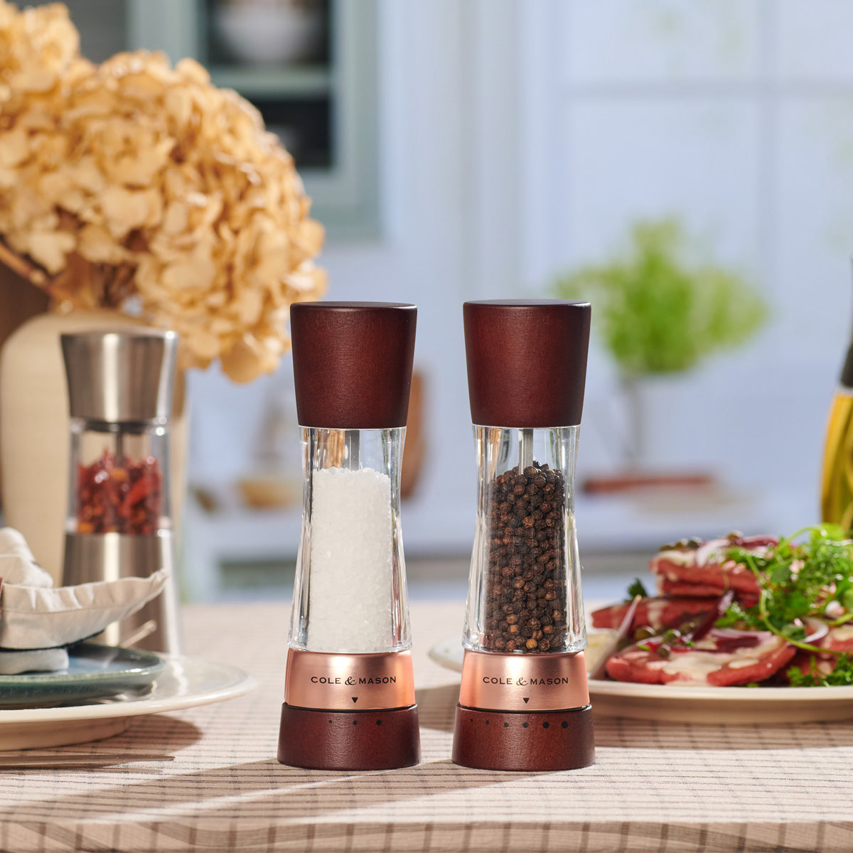 Two Cole & Mason salt and pepper grinders with dark wood tops and bases sit on a table, with food and flowers blurred in the background. The left grinder contains salt, and the right contains peppercorns.