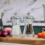 The Cole & Mason UK Everyday Salt & Pepper Mill Set Acrylic 110mm features two mills with adjustable ceramic mechanisms, filled with salt and black peppercorns, displayed on a wooden board amid fresh vegetables in a kitchen.