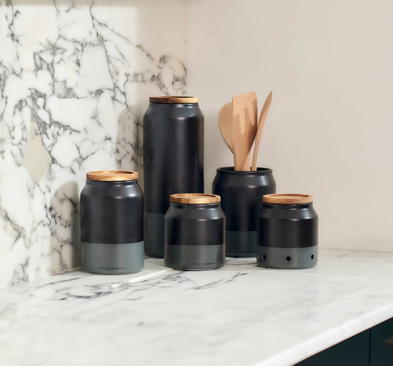 Five matte black and gray kitchen canisters with wooden lids are arranged on a white marble countertop. One canister holds wooden utensils. The background features white marble with gray veining.