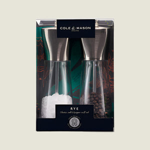 The Cole & Mason UK Rye Salt & Pepper Mill Set 160mm features clear bodies with metal tops and durable ceramic mechanisms, filled with coarse salt and whole peppercorns, shown against a green background.