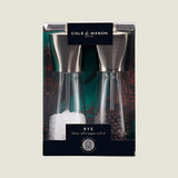 The Cole & Mason UK Rye Salt & Pepper Mill Set 160mm features clear bodies with metal tops and durable ceramic mechanisms, filled with coarse salt and whole peppercorns, shown against a green background.