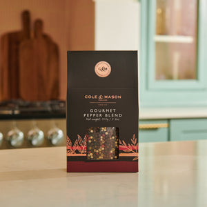 A box of Cole & Mason Gourmet Pepper Box sits on a kitchen counter, with a stove and blue cabinets behind it. A clear window reveals colorful peppercorns inside, ideal for grinding fresh flavor into your favorite dishes.