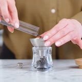 A person pours whole peppercorns into a glass pepper grinder, using a metal funnel, on a white marble countertop. The person is wearing a brown shirt.