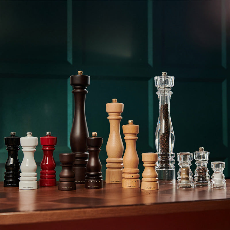 A variety of pepper and salt mills in different sizes, colors, and materials—black, white, red, natural wood, and clear acrylic—are displayed on a wooden surface against a dark green background.