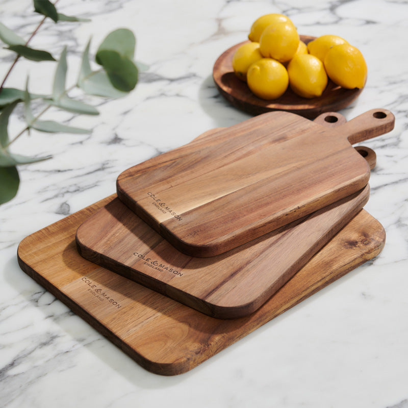 Three wooden cutting boards of different sizes are stacked on a white marble countertop. In the background, a wooden bowl holds several yellow lemons, and green foliage is partially visible to the side.