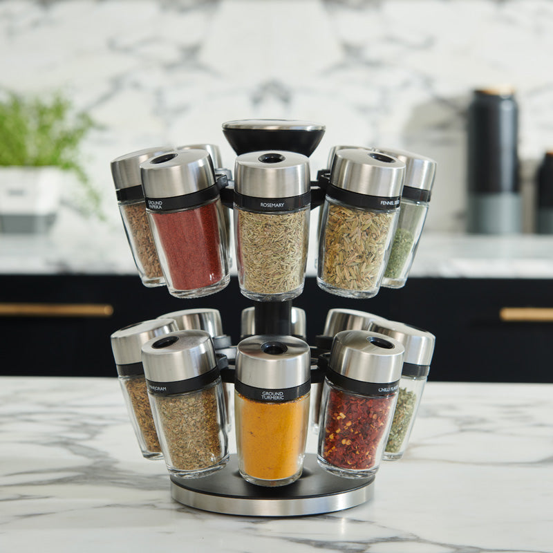 A rotating spice rack with twelve glass jars filled with various colorful spices, placed on a marble kitchen countertop. Each jar has a metal lid and a label indicating the spice inside.