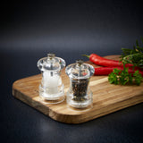 #85mm#pepper
Two Cole & Mason Acrylic Capstan Salt & Pepper Mills with a clear design rest on a wooden board, surrounded by red chili peppers, parsley, and rosemary against a dark background.