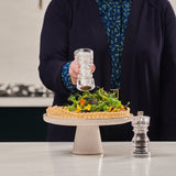 A person sprinkles pepper from the Cole & Mason London Acrylic Salt Shaker & Pepper Mill Set onto a tart with greens and edible flowers on a pedestal stand, while the modern set completes the stylish tableware display.