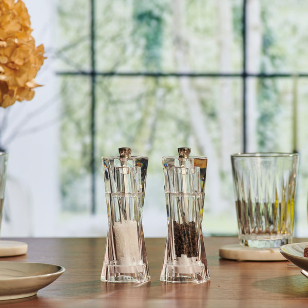 The Cole & Mason Aldeburgh Salt & Pepper Mill Set, featuring clear glass and premium grinders, sits on a wooden table amid plates, glasses, and a vase of dried flowers, with a large window and blurred outdoor view behind.