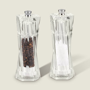 The Cole & Mason Aldeburgh Salt & Pepper Mill Set features two clear acrylic mills with adjustable grinding, filled with black peppercorns and coarse sea salt, standing side by side on a light background.