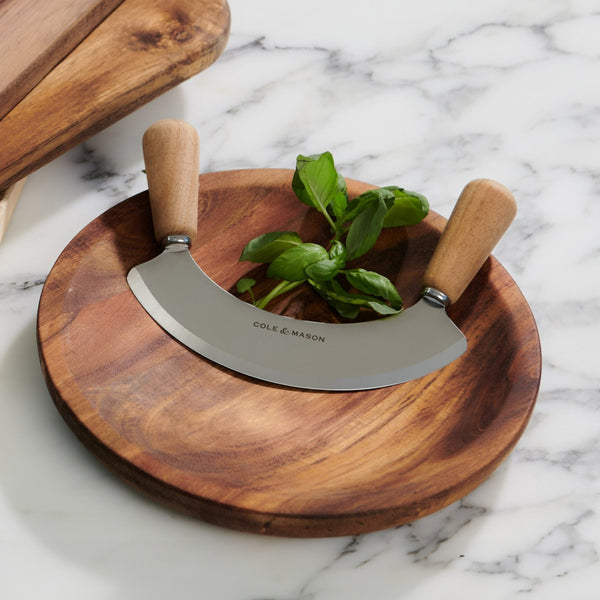 The Cole & Mason Ashden Mezzaluna and Board, featuring wooden handles and a round cutting board with fresh basil leaves, is ideal for food prep and looks elegant on a white marble countertop.