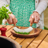 Wearing a green patterned dress, a person uses the Cole & Mason Ashden Mezzaluna and Board to chop fresh herbs outdoors. A red pepper grinder and herb sprig nearby add charm to the rustic food prep scene.