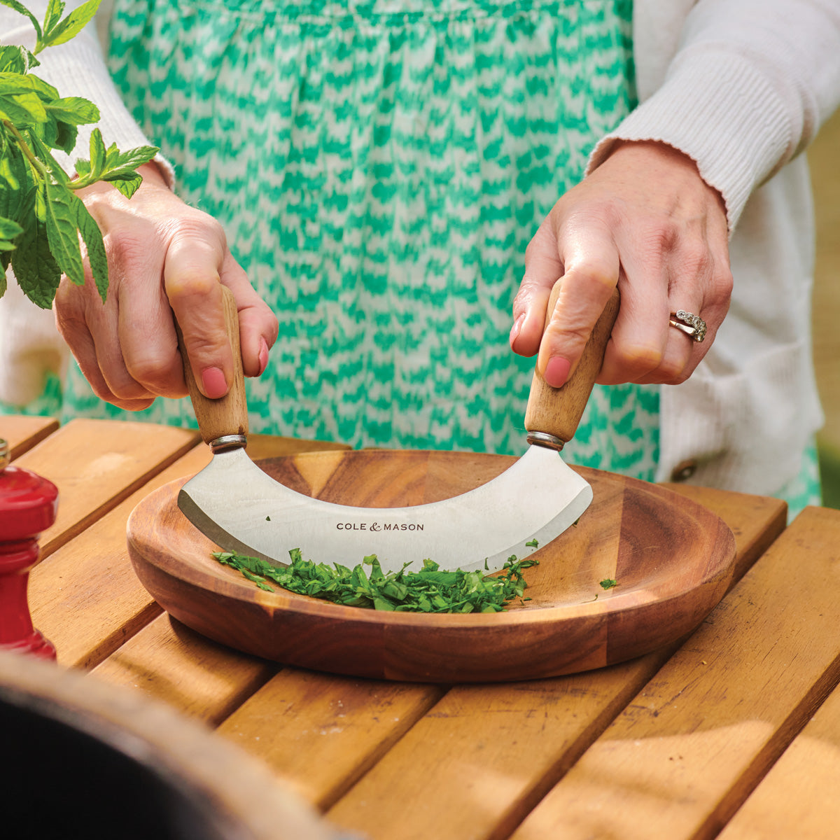 A person uses a double-handled herb chopper to finely cut fresh herbs on a round wooden plate. The individual wears a green patterned dress and a white cardigan. A plant and a red item are nearby on the wooden table.