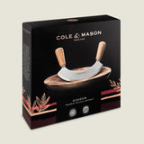 The Cole & Mason Ashden Mezzaluna and Board set features a curved chopping tool with wooden handles on an acacia wood board, displayed in stylish packaging with gold and red accents. Ideal for quick, efficient food prep.