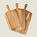 The Cole & Mason 3 Piece Barkway Board Bundle features three rectangular acacia chopping boards with handles, arranged overlapping to showcase their premium wood grain—each stamped with the brand name for a stylish kitchen essential.