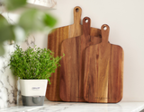 Two Cole & Mason Barkway Boards stand upright on a marble countertop beside a potted green herb. Crafted from premium acacia wood, this 2 Piece Board Bundle is an essential kitchen addition for any home chef.