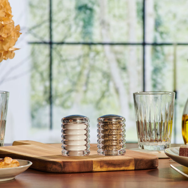 The Cole & Mason Beehive Shakers Set, featuring two durable acrylic shakers for salt and pepper, sits on a wooden board amid glasses and plates on a dining table, with blurred greenery visible through a window in the background.