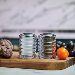 The Cole & Mason Beehive Shakers Set, made of durable acrylic, rests on a wooden board with an artichoke, yellow cherry tomato, and dark grapes, all set against a softly blurred background.
