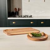 The Cole & Mason Berden Carving Board & Ashden Mezzaluna Bundle rests on a light kitchen counter with fresh green herbs. In the background, dark cabinets and a stovetop complete the scene.